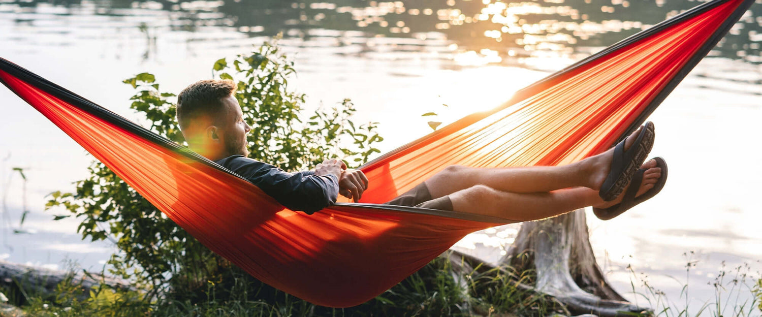 Man relaxing in a hammock wearing comfortable men's sandals by the river during sunset.