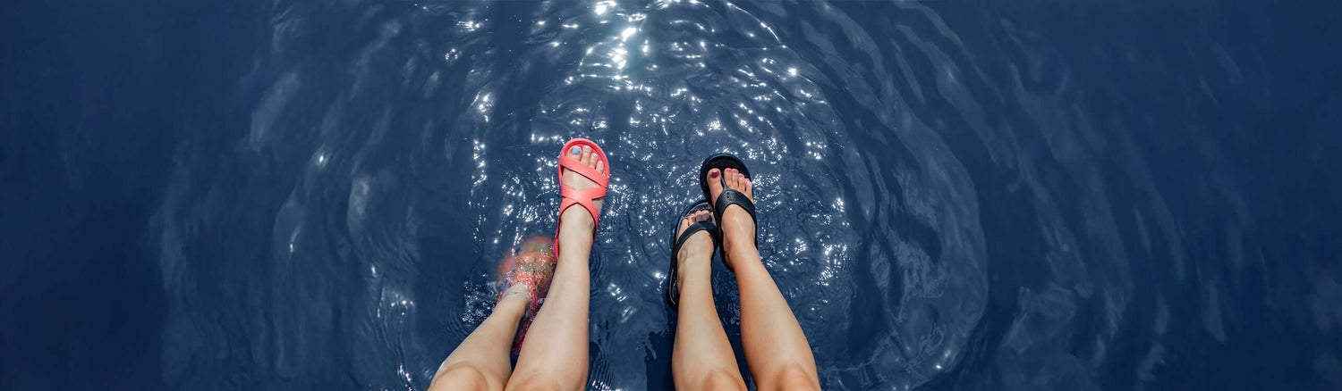 Two pairs of feet in comfortable women’s sandals splashing in water during a sunny day.