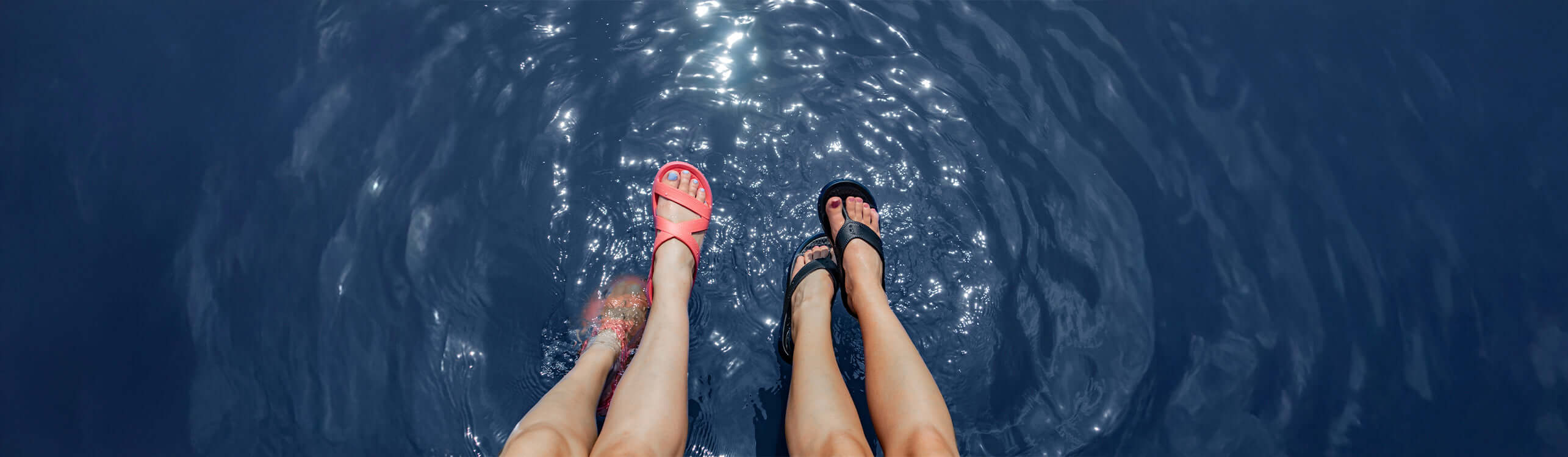 Two pairs of feet in comfortable women’s sandals splashing in water during a sunny day.