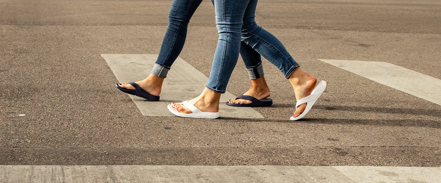 two women crossing a street intersection, legs and feet only visible, wearing jeans and cascade flip flops in white water and eclipse black