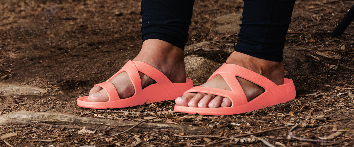 women's feet wearing the nuusol hailey slide in coral sunrise color. the picture was taken in a forest, and you can see the twigs and dirt of the forest floor.